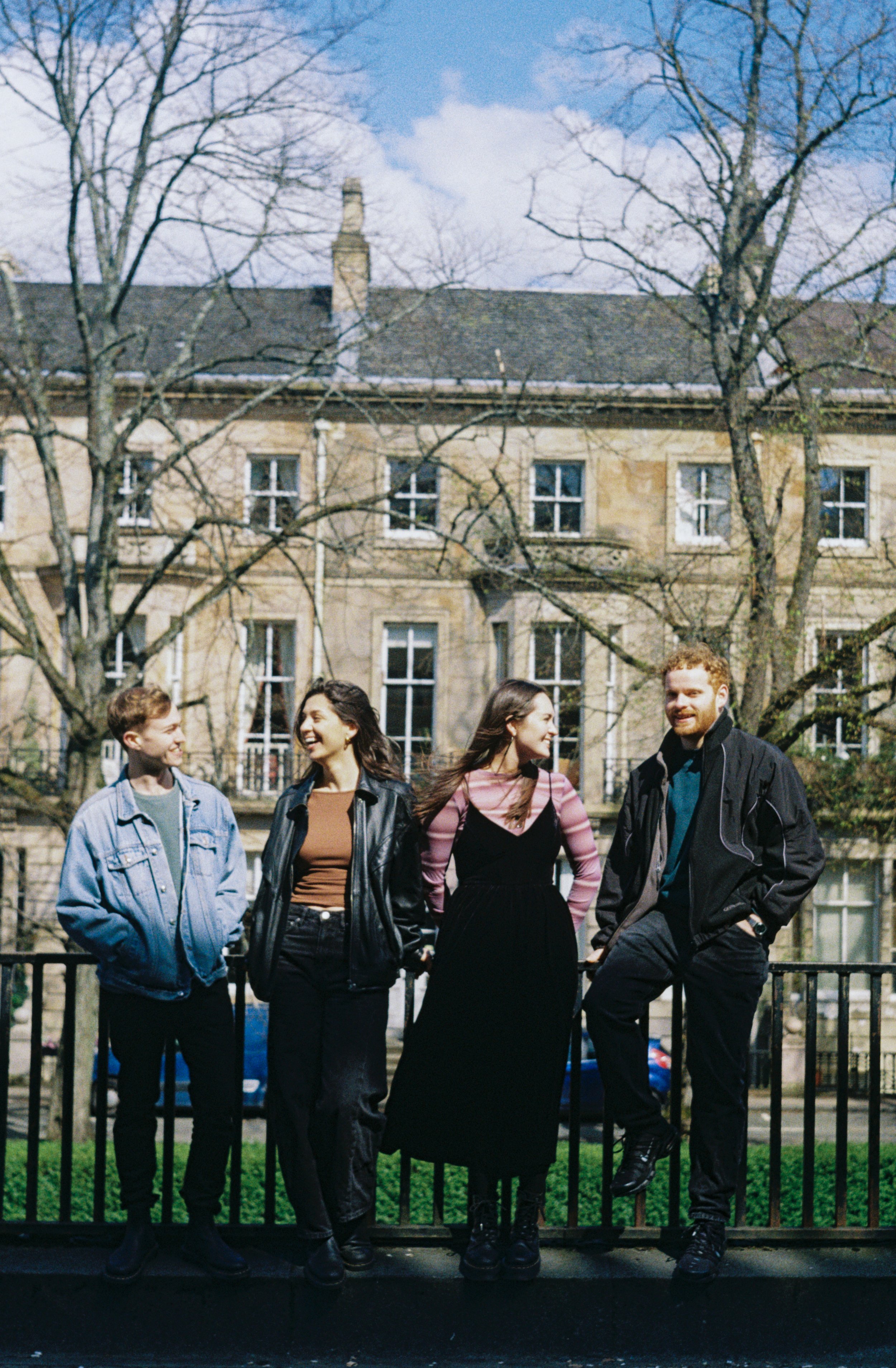 Five young adults standing outdoors behind a black fence, smiling and talking to each other. Two women and three men, dressed casually, with a backdrop of leafless trees and old brick buildings under a blue sky.