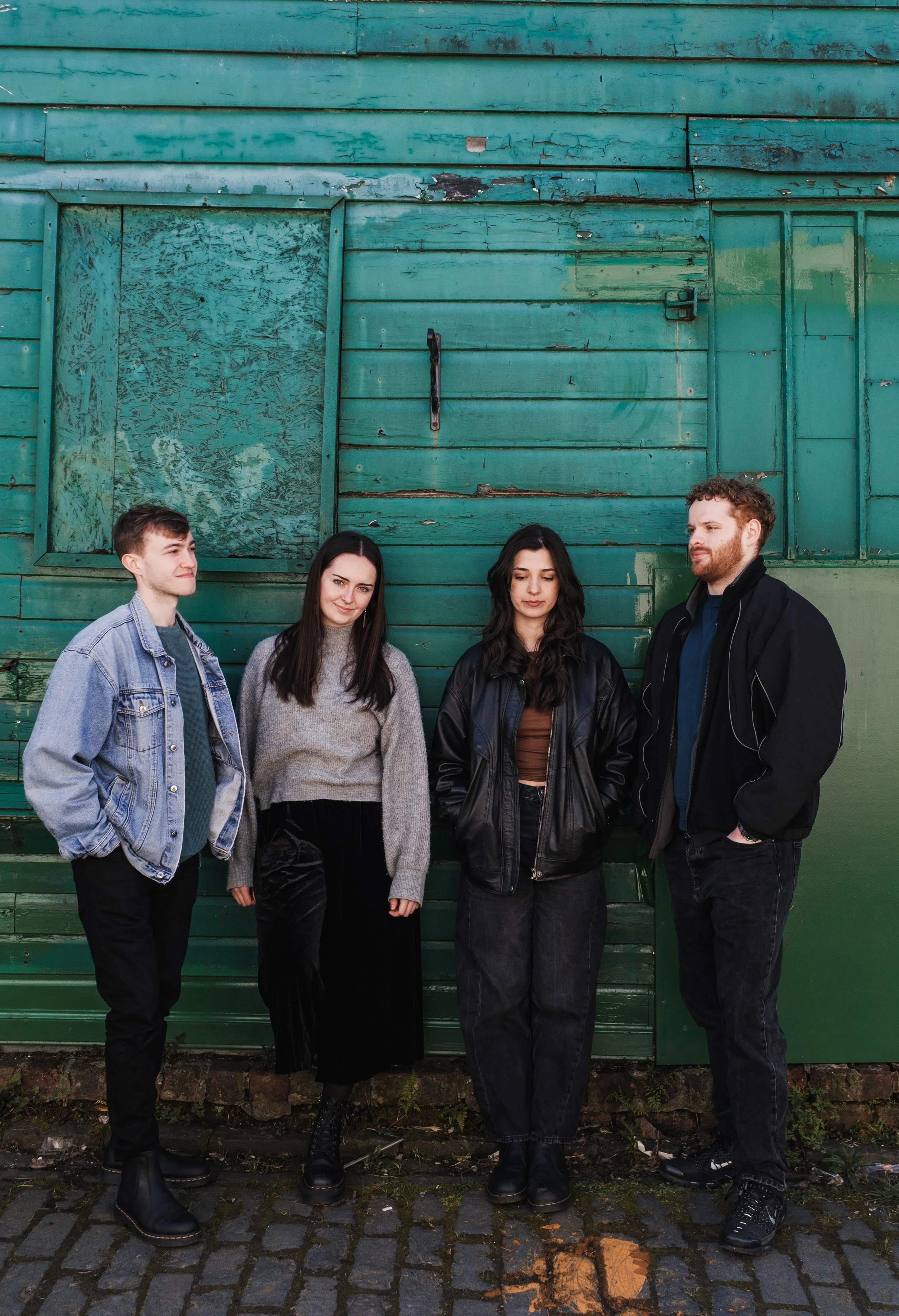 Group of four young adults standing against a weathered green wooden wall with a boarded-up window. They are dressed in casual, dark clothing and have varied hairstyles.