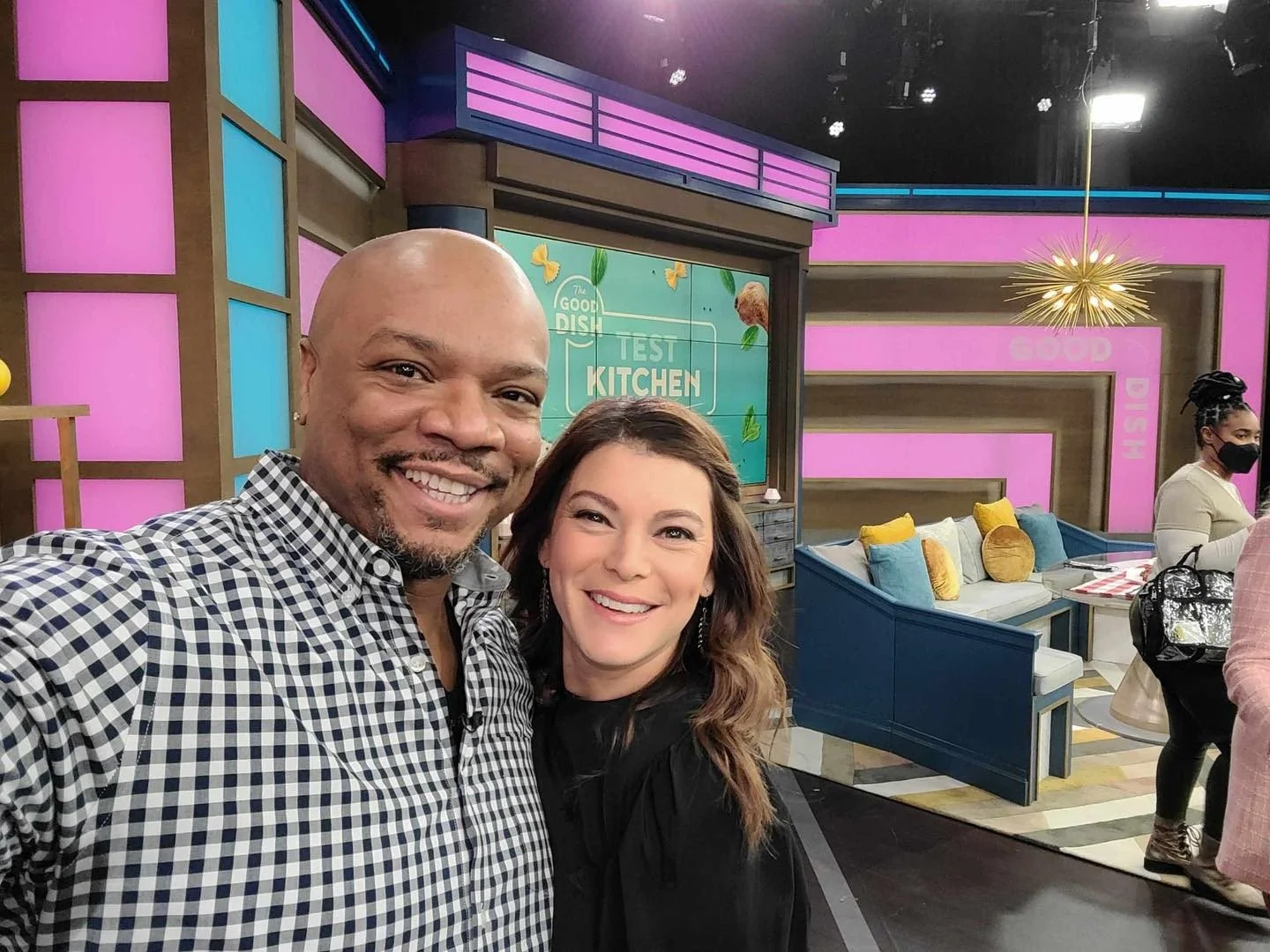 Two people taking a selfie on a TV studio set for a show called "The Good Dish," featuring colorful backgrounds and a seating area. Another person can be seen in the background wearing a face mask.