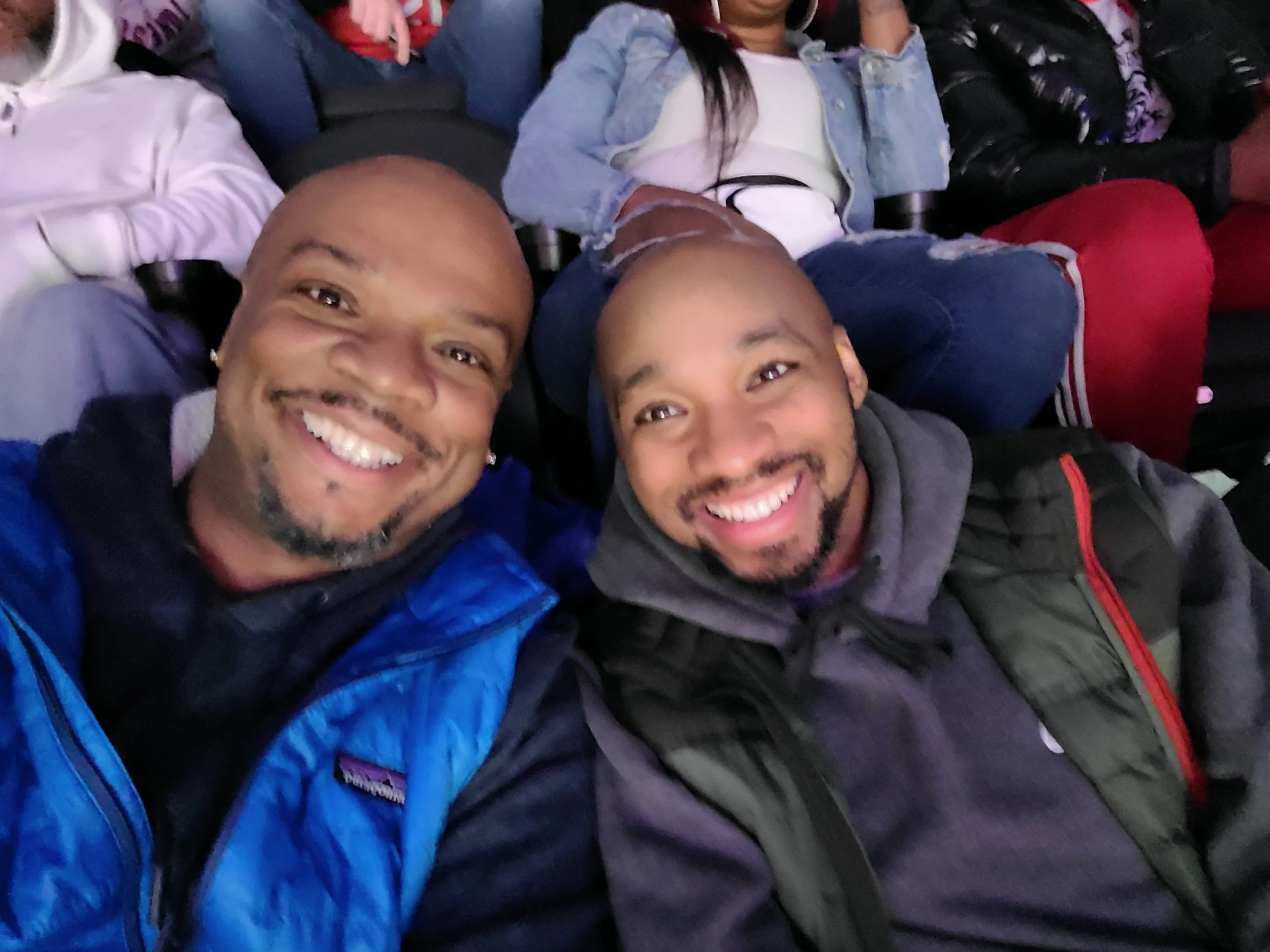 Two smiling people taking a selfie in a crowded seating area, wearing casual jackets and hoodies.