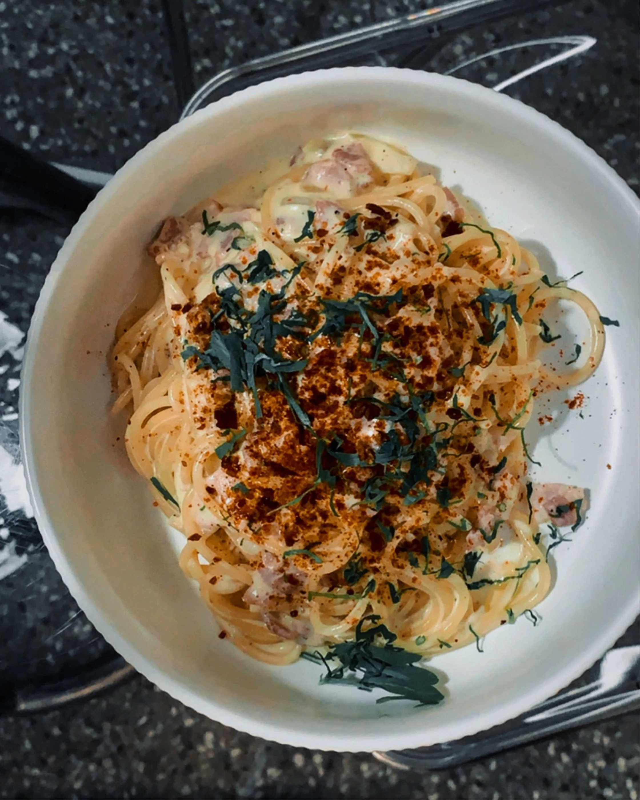 Spaghetti carbonara in a white bowl, topped with parsley and paprika.
