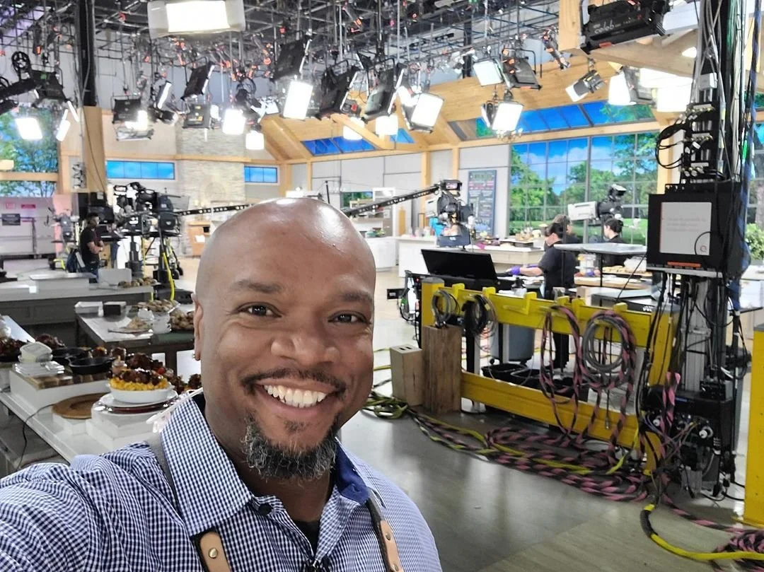 Smiling man taking a selfie on a bright TV studio set with cameras and lighting equipment.
