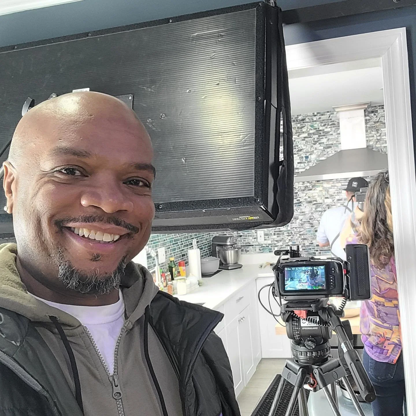 Man taking a selfie in a kitchen with professional camera and crew filming in the background.