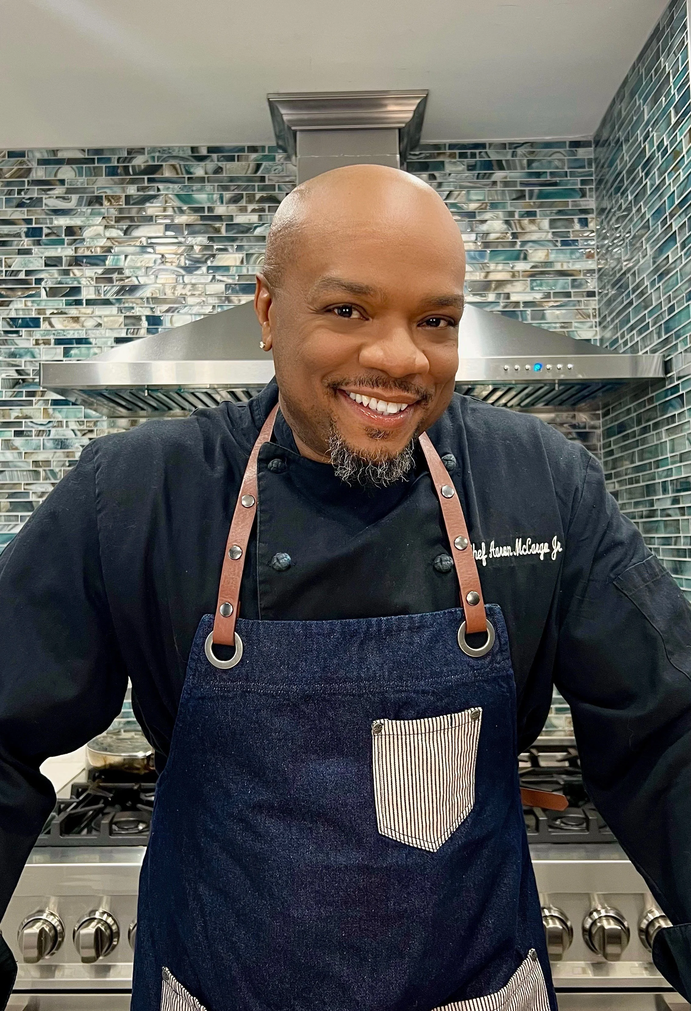 A smiling person in a chef's uniform poses in a kitchen with a modern stove and blue mosaic tile backsplash.