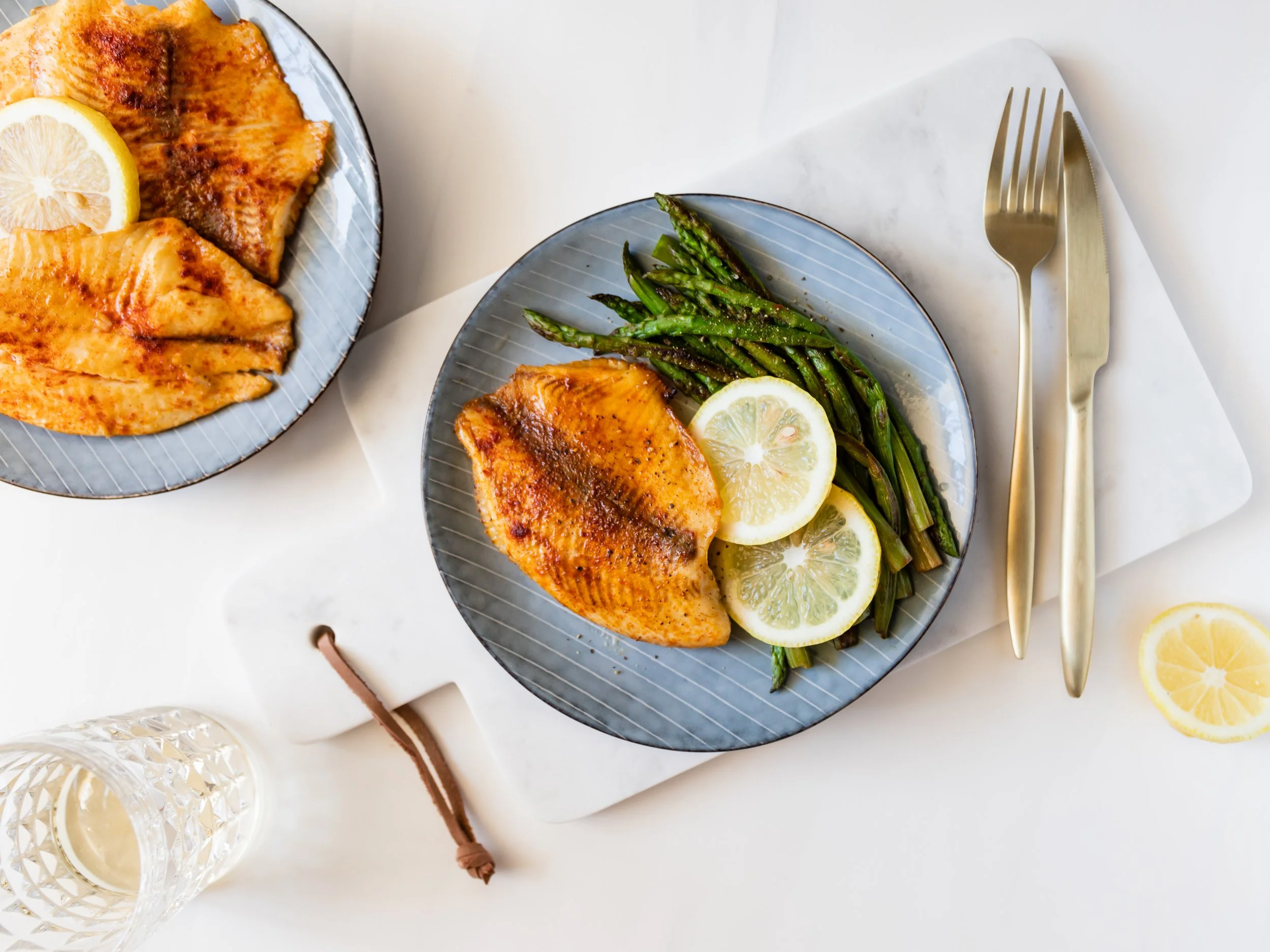Grilled fish fillet with lemon slices and asparagus on a blue plate, set on a white marble board, with a fork and knife beside it, accompanied by a drinking glass.