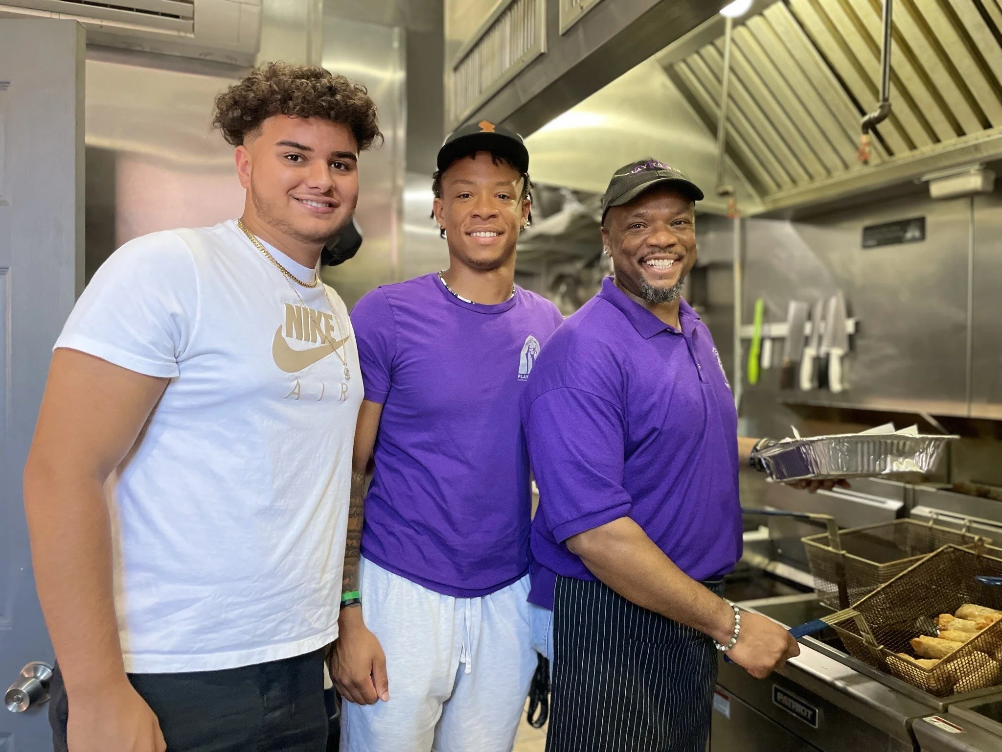 Three men standing in a commercial kitchen, smiling, with one holding a tray next to a fryer filled with food.