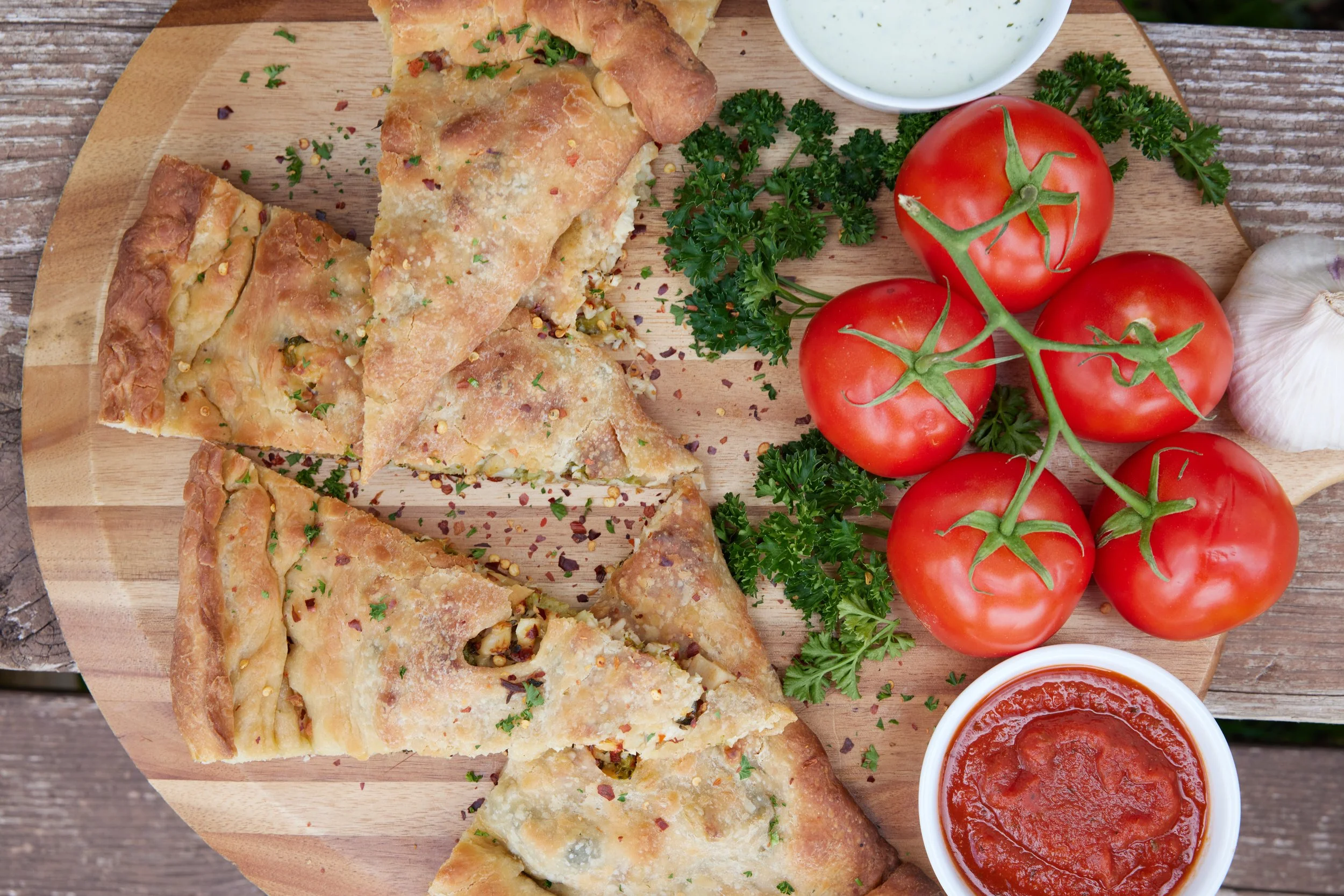 Assorted calzone slices on a wooden board with fresh tomatoes, parsley, garlic, and a cup of marinara sauce.