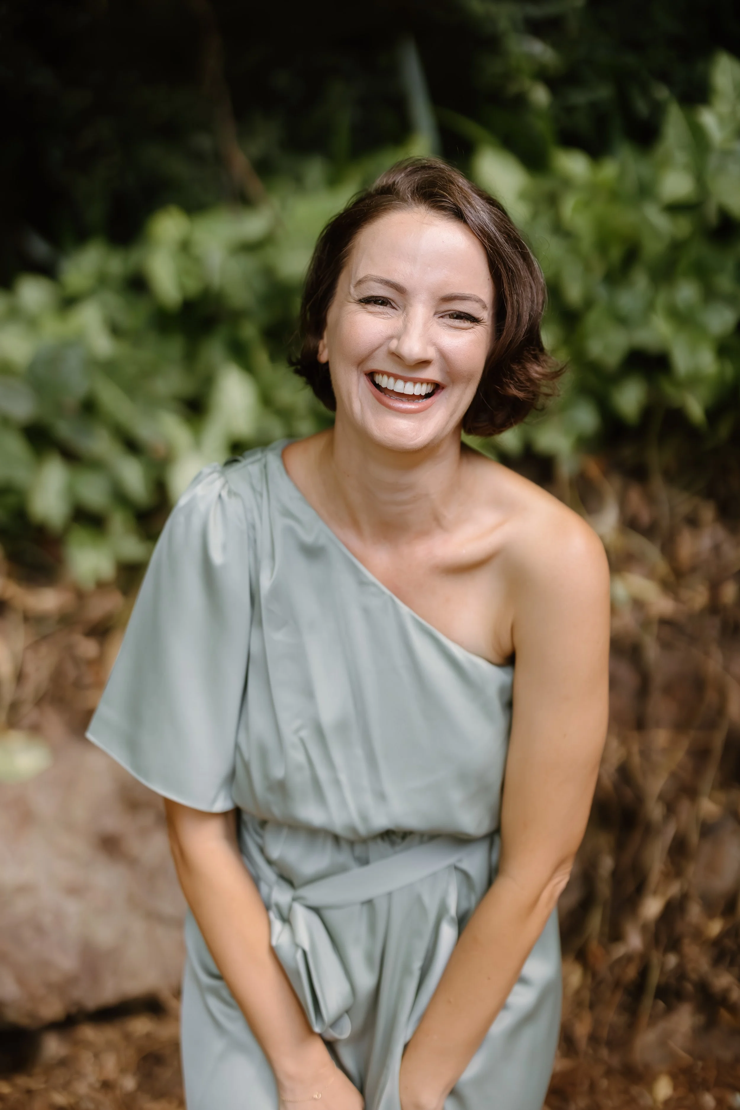 Three smiling people, two women and one man, posing together at a social event or gathering. The man is wearing a black shirt and tie, the woman in the middle is wearing a white dress, and the woman on the right is wearing a light-colored dress with green earrings.