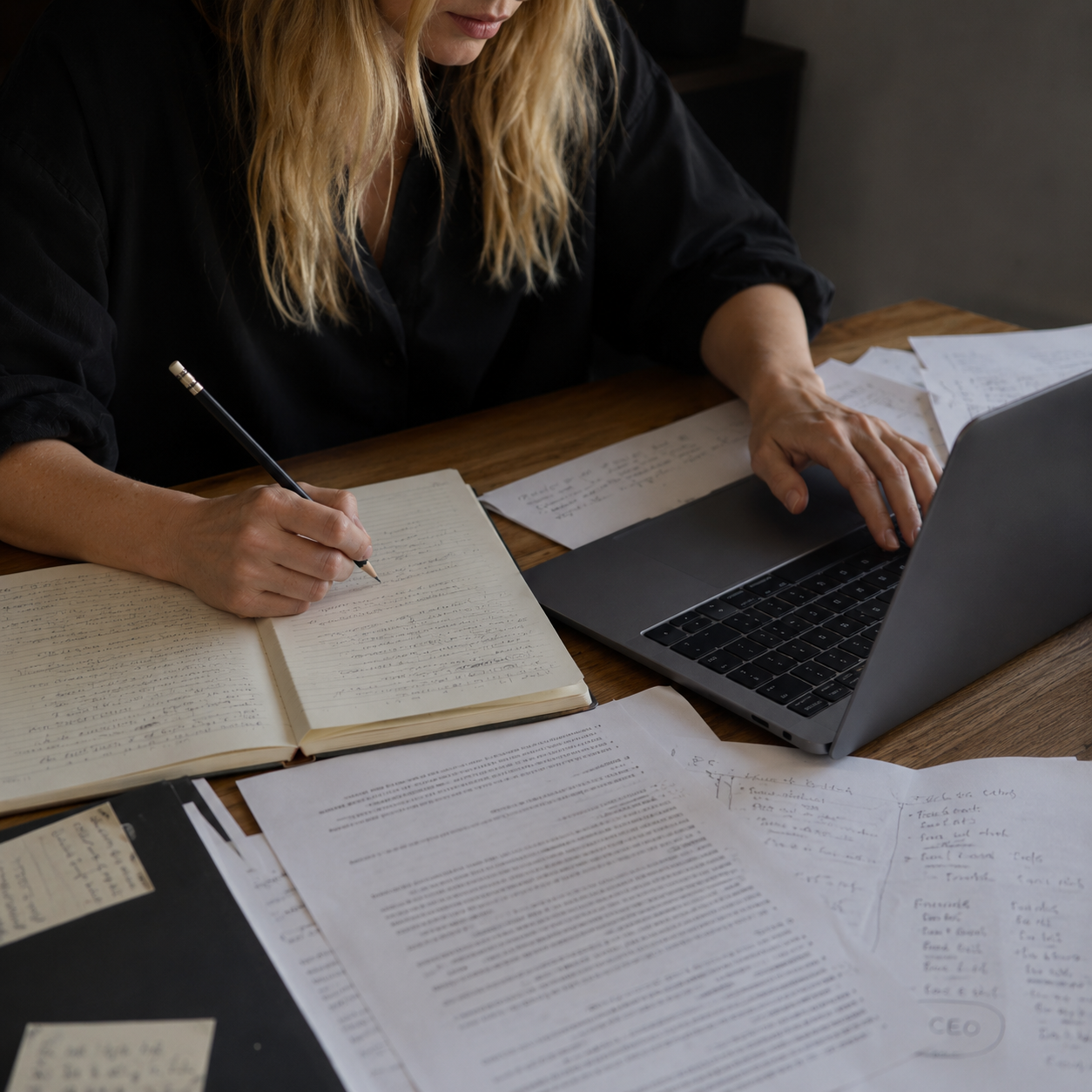 a blonde woman writing on her macbook, paper scattered everywhere