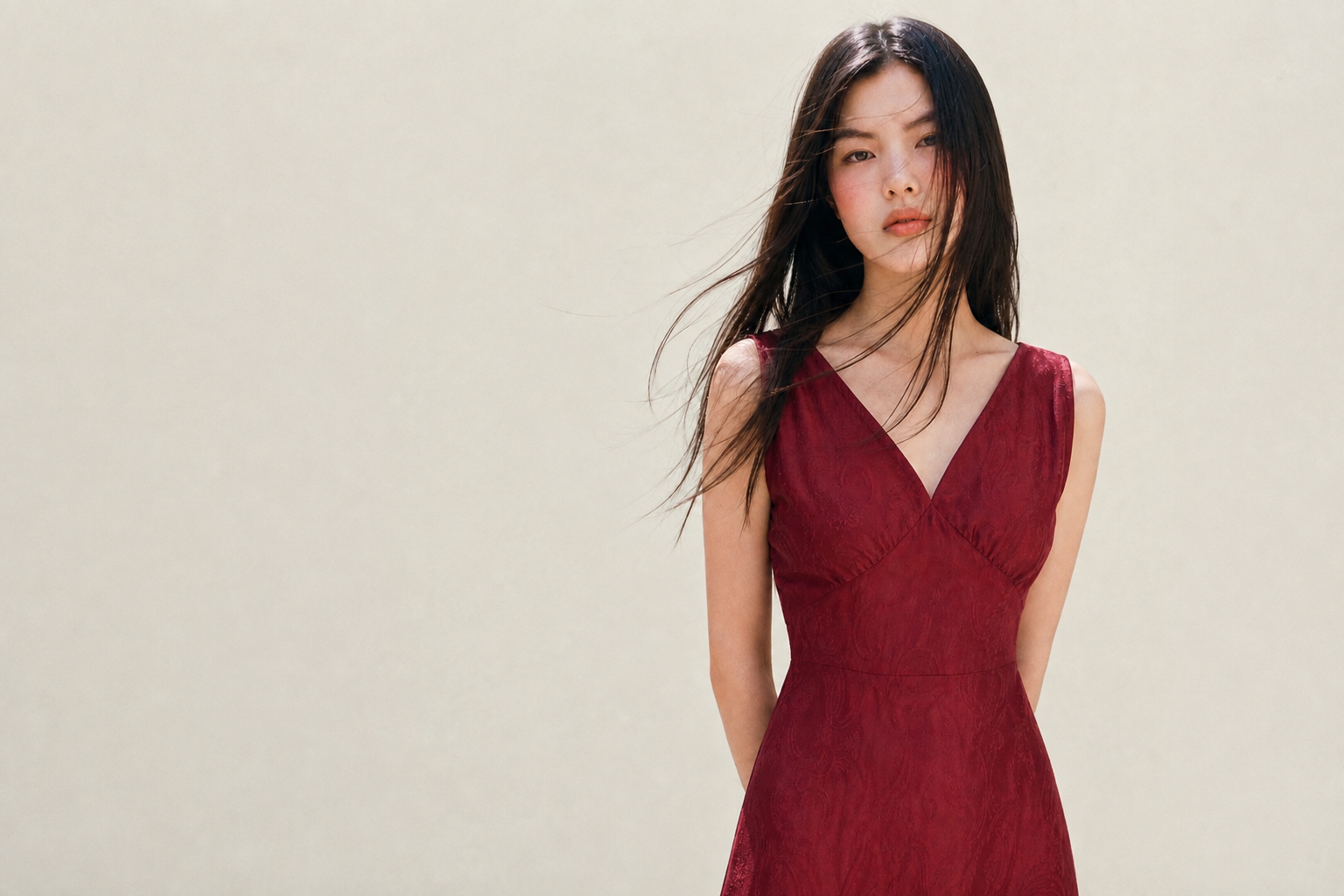 A woman with long dark hair in a sleeveless red dress standing against a plain light background.