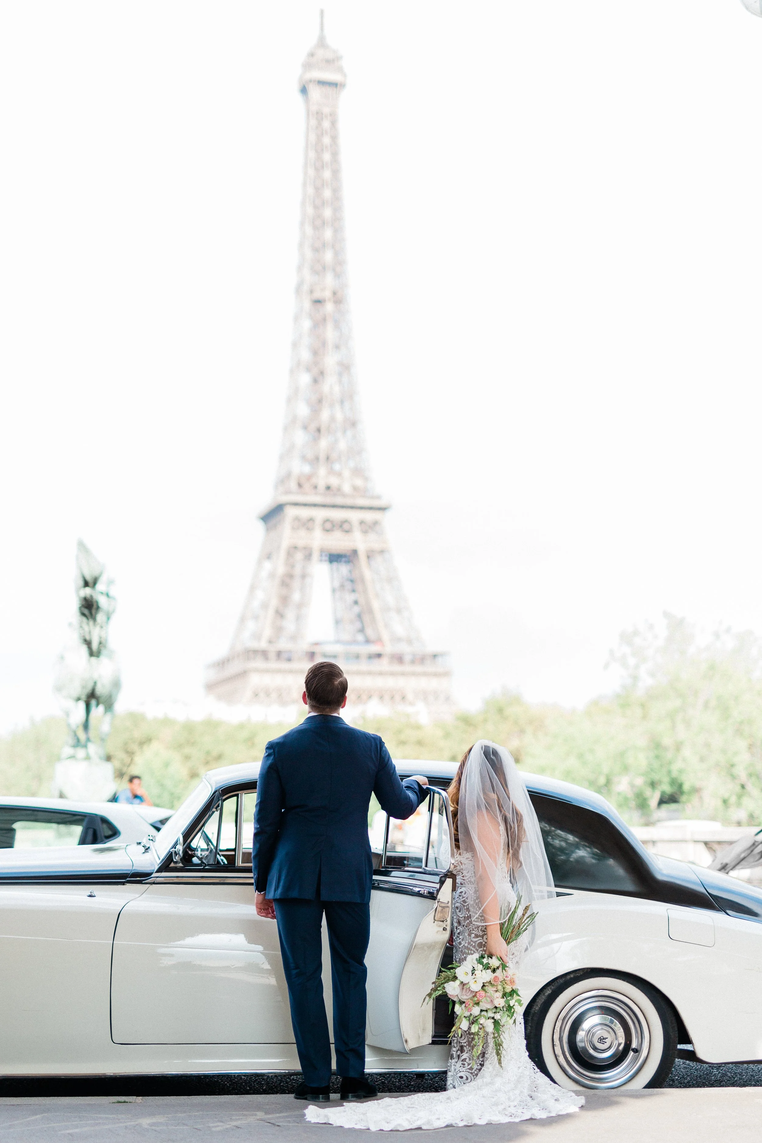 A bride and groom standing beside a vintage white car in front of the Eiffel Tower, with the groom holding the car door open and the bride holding a bouquet, during a wedding photo shoot.