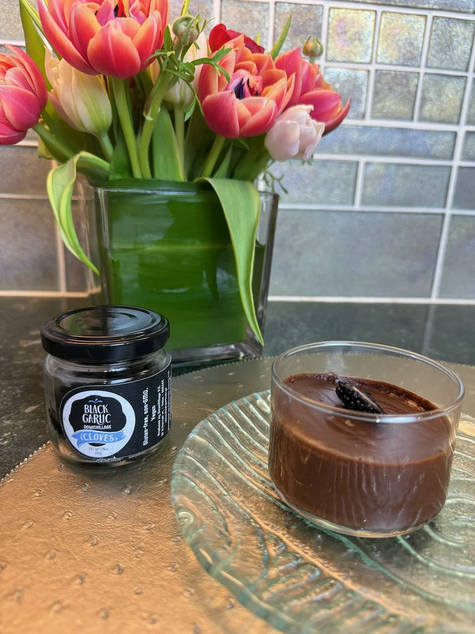 A romantic still life of very dark chocolate mousse, styled with fresh flowers and a jar of black garlic cloves..