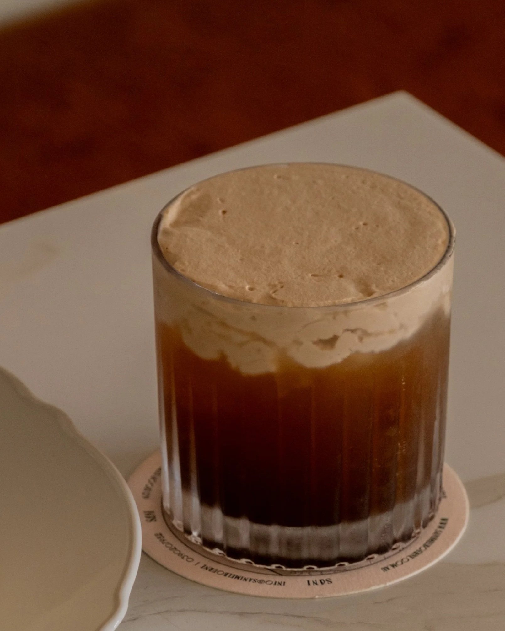 A glass of iced coffee with a layer of whipped cream on top, placed on a round coaster on a white surface.