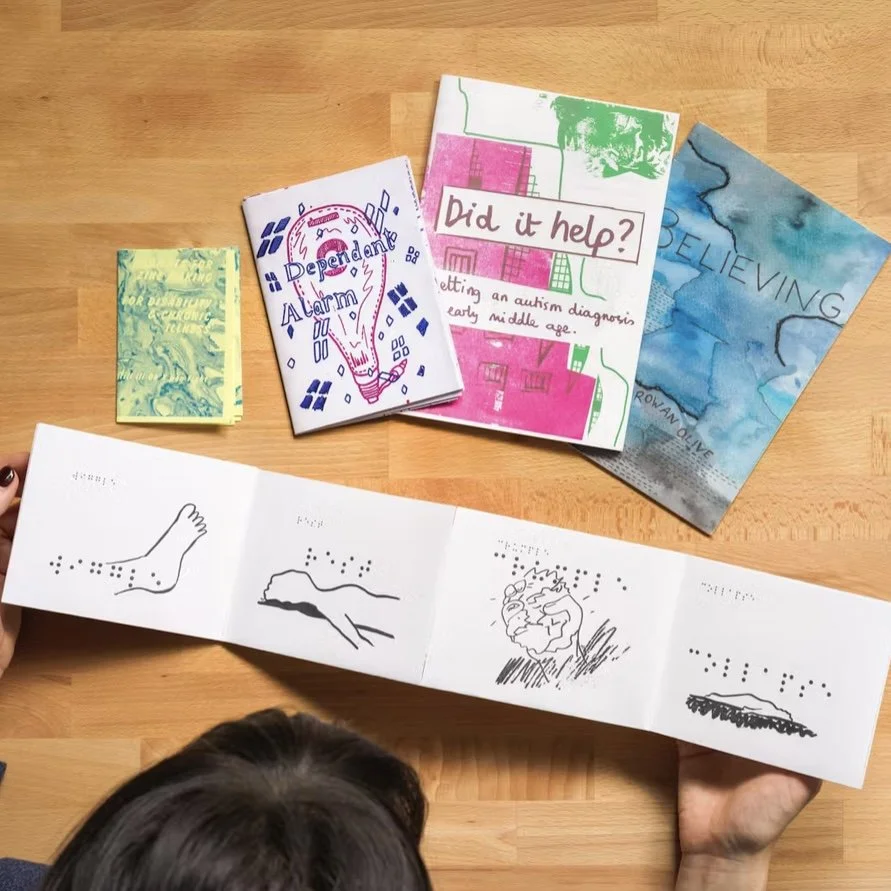 Photo of a person reading Embodied zine, which is spread out on a table. The perspective is looking down at the table, so the top of the person's head and hands are shown but the zine is the center. Four other zines are on the table nearby.