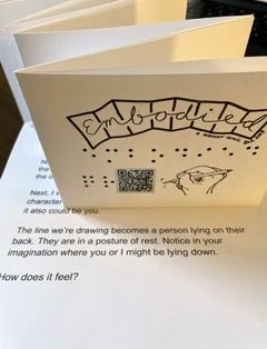 Photo of Embodied accordion book showing the title page, resting on top of a page of text from the inside of the folded art book.