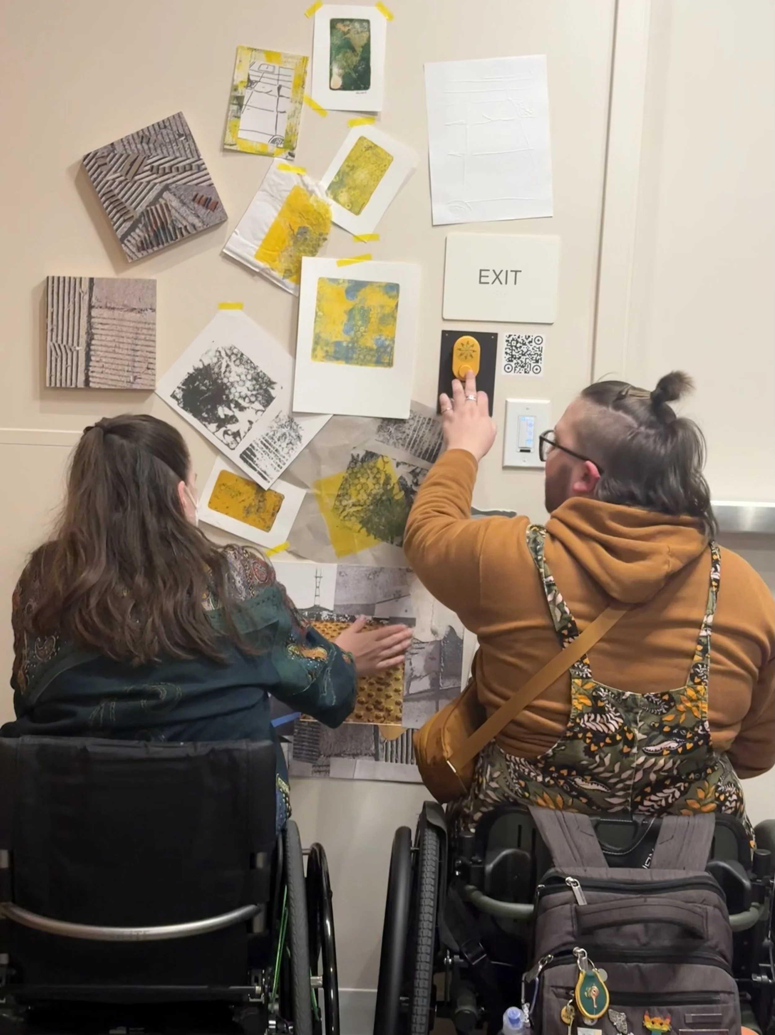 Two visitors who are wheelchair users with their backs to the camera are touching “Bubbling Up, “ an wall assemblage.