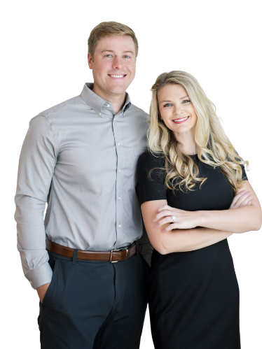 A young man and woman smiling, standing close together against a plain background. The man is wearing a light gray long-sleeve button-up shirt and dark pants, with his hands in his pockets. The woman is wearing a black short-sleeve top and dark pants, with her arms crossed. Both are looking at the camera with friendly expressions.