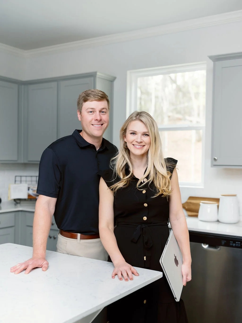 A man and woman standing in a modern kitchen, smiling at the camera. The woman is holding a laptop, and the man is resting his hand on the kitchen counter.