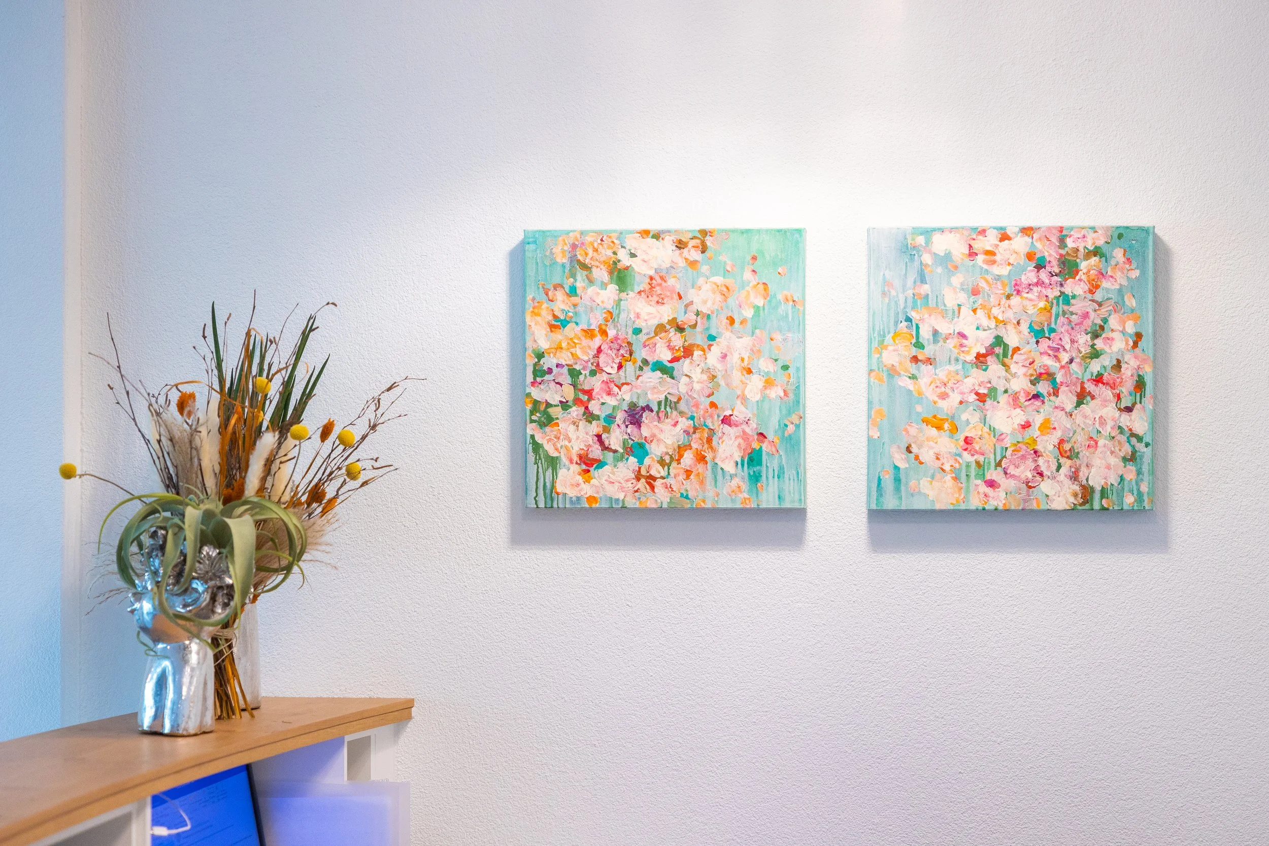 Innenraum mit zwei bunten Blumenbildern an der Wand und einem dekorativen Blumenarrangement in einer silbernen Vase auf einem Regal.