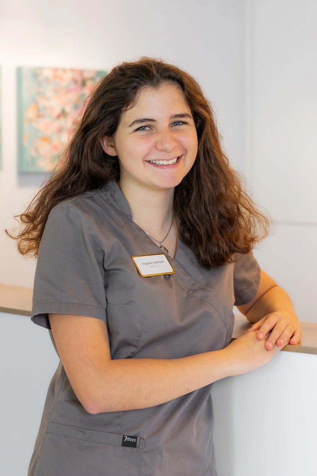 Junge Frau in grauem medizinischem Kittel lächelt in die Kamera, während sie sich an einen Empfangstresen lehnt.