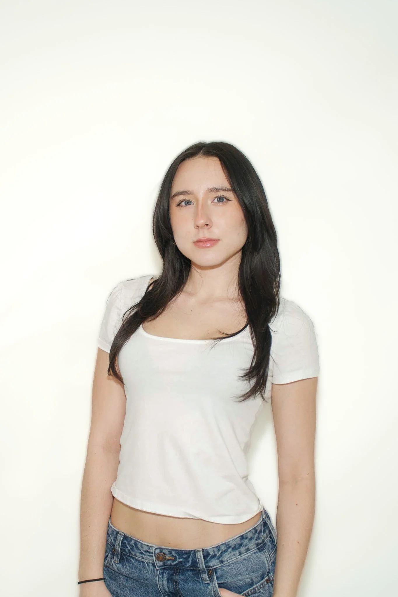 A young woman with long dark hair and fair skin, wearing a white crop top and blue jeans, standing against a plain light background.