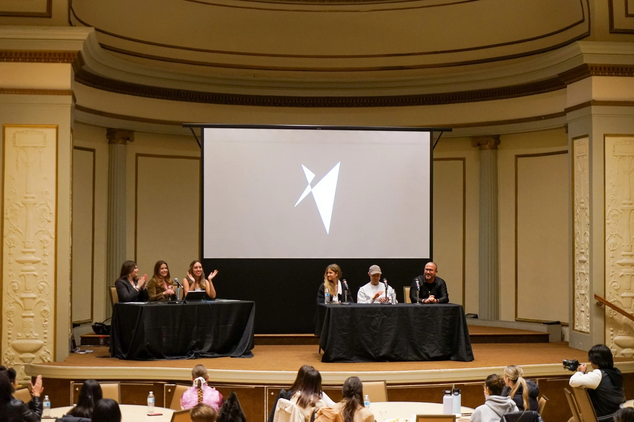 A panel discussion with six people seated at two tables on a stage in an ornate room, with an audience watching and a large screen displaying a stylized geometric logo behind them.
