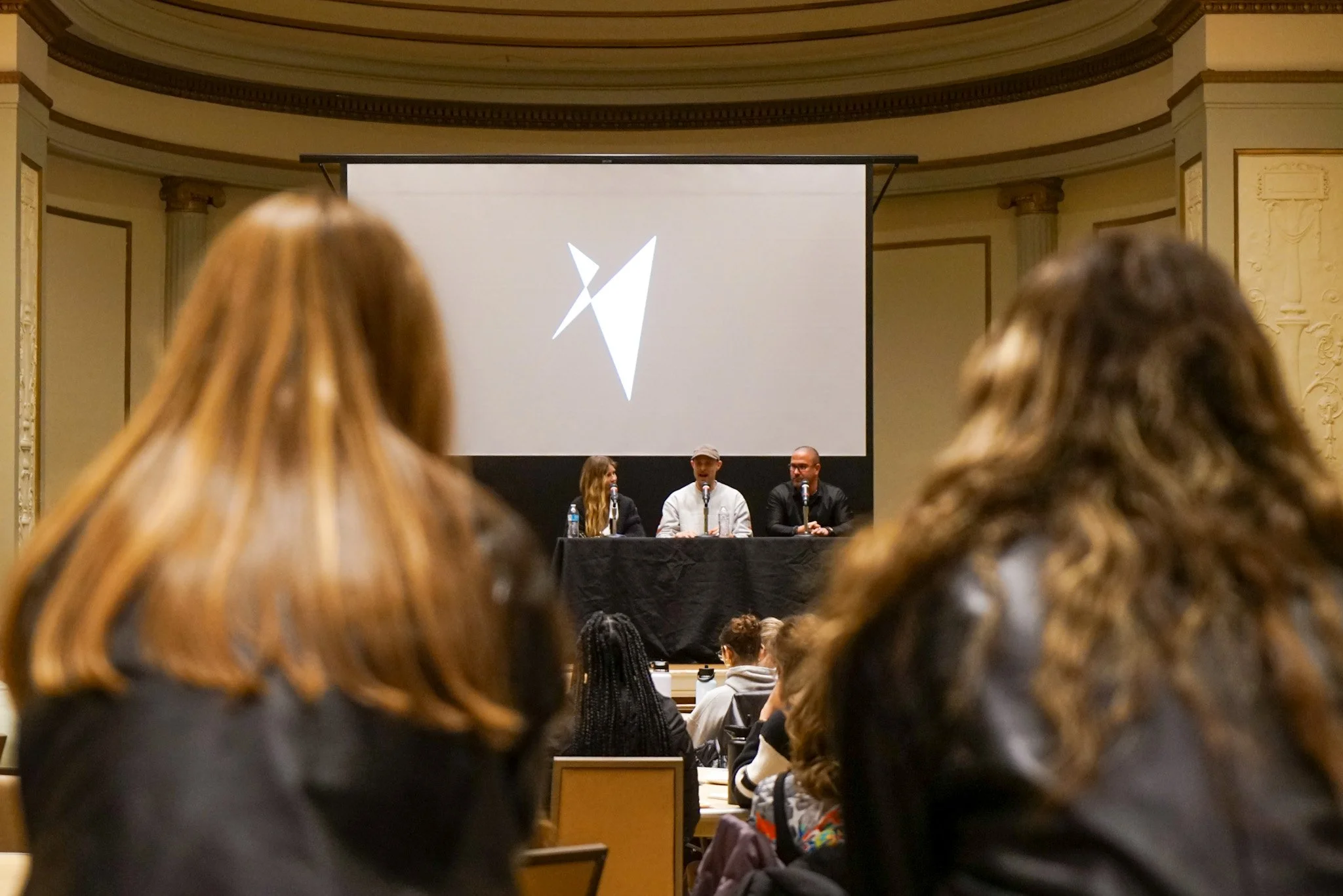 Panel discussion at conference with three speakers sitting at a table in front of a large projection screen, audience members seated facing them.