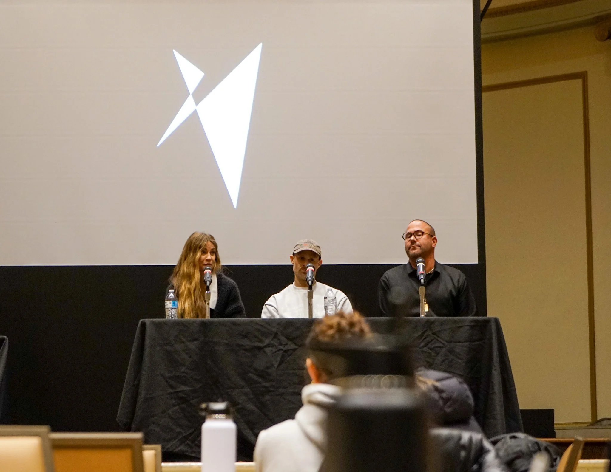 Three people sitting at a panel in front of a large screen with a white abstract logo. The panel includes a woman with long wavy hair, a man in a cap, and a man with glasses. They are speaking into microphones.