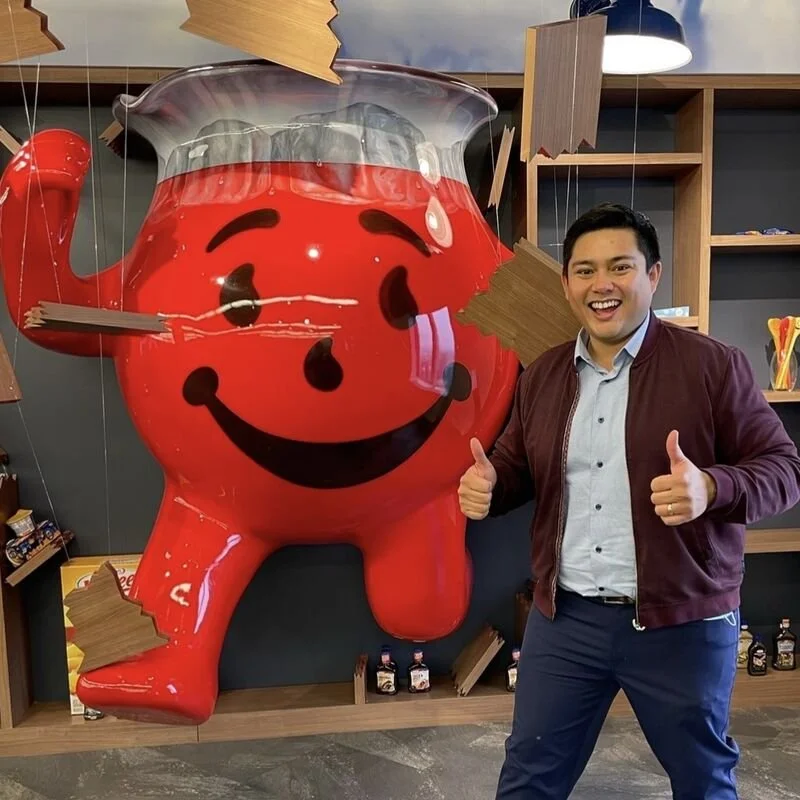 A man standing next to a large, inflatable red character with a smiling face, in what appears to be an indoor setting with shelves and decorations.