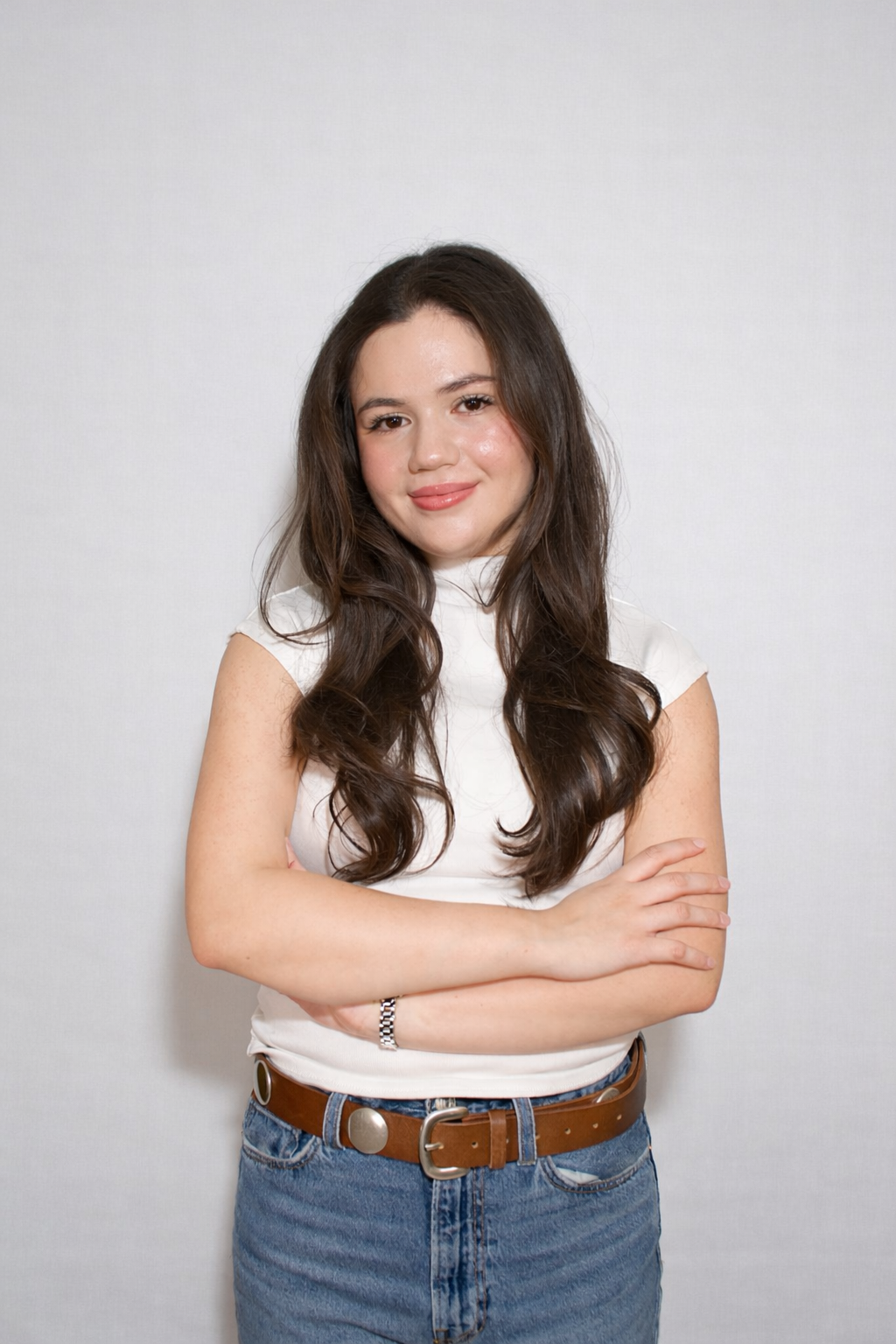 A young woman with long brown hair, wearing a white sleeveless top and blue jeans, standing against a plain light gray background with her arms crossed and smiling.