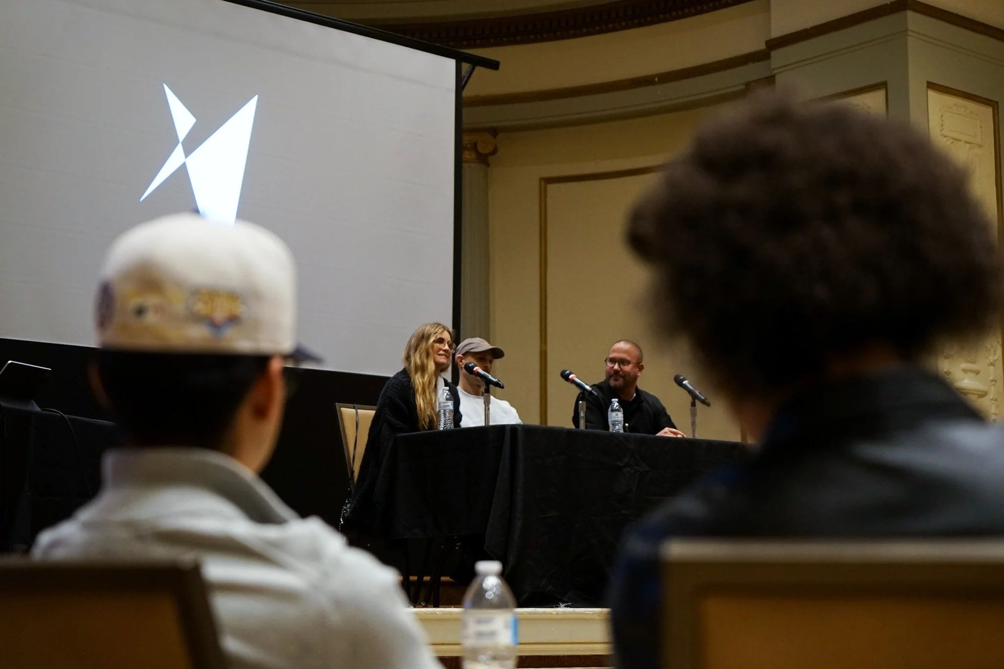 Panel discussion at a conference with three speakers seated at a table in front of an audience, with a large projection screen behind them.