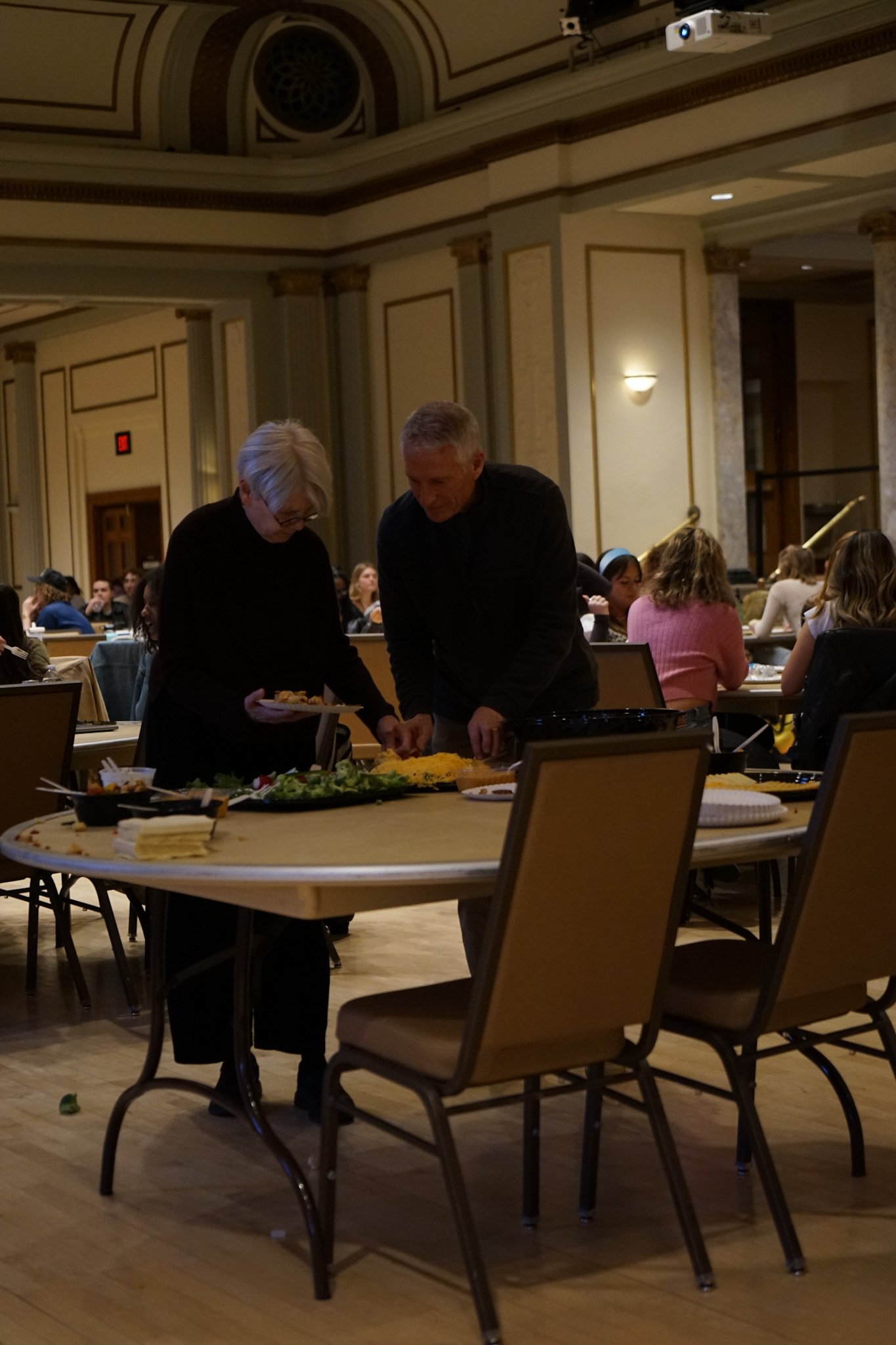 Two people serving themselves food from a buffet table in a banquet hall with many seated guests in the background.