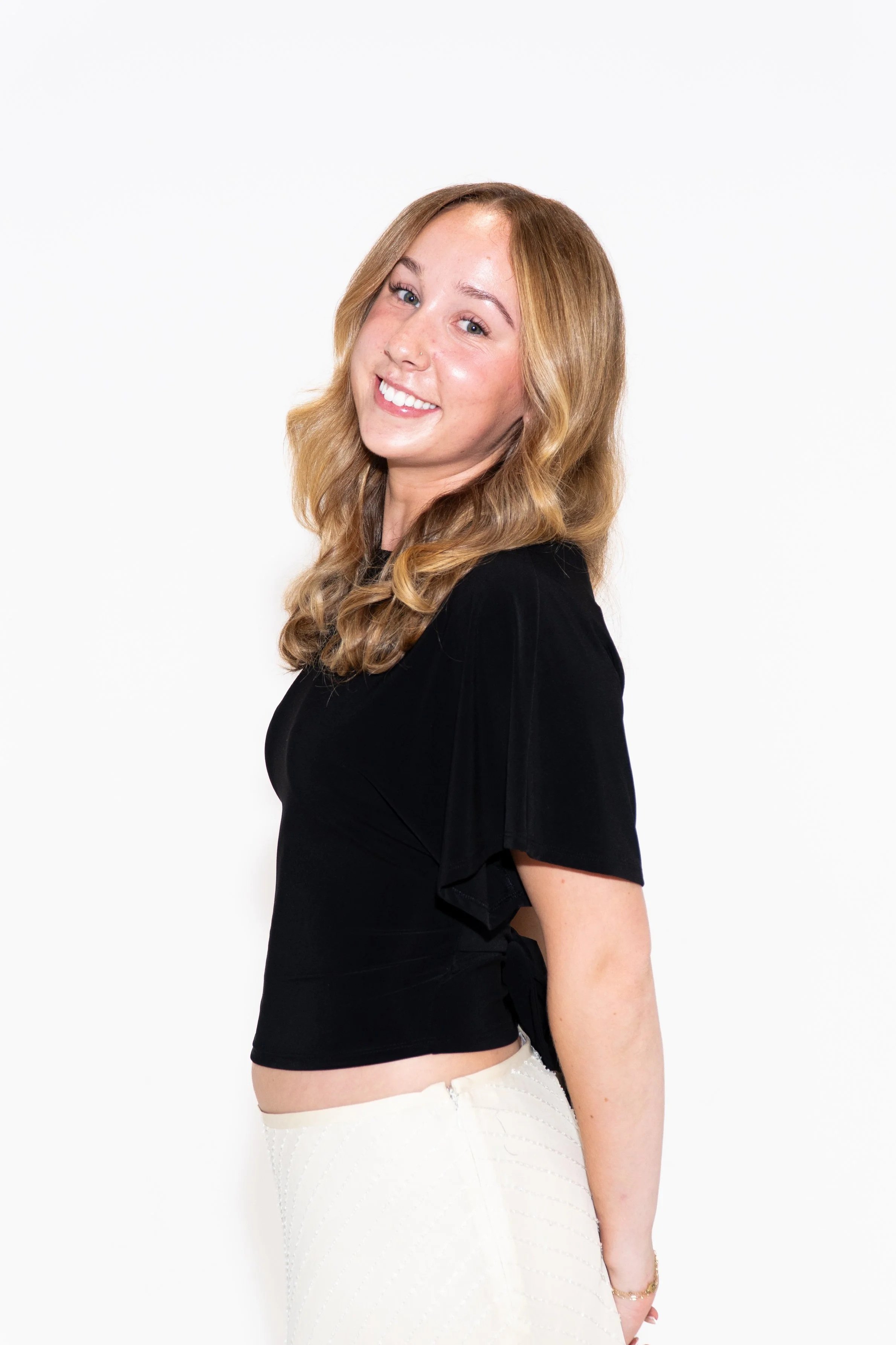 A young woman with long, wavy brown hair wearing a white t-shirt and jeans, standing against a plain white background, with a confident expression, hands on her hips, and jewelry including a necklace, ring, and bracelet.