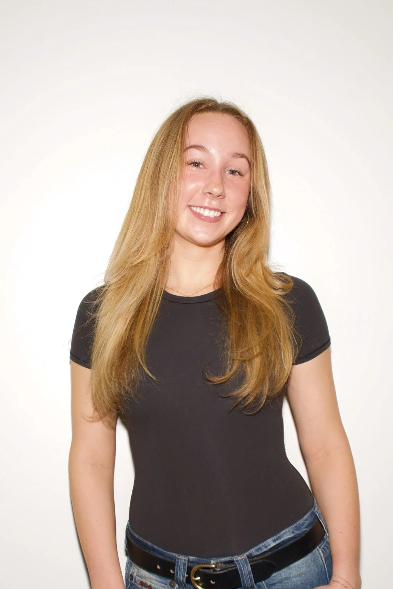 A young woman with long red hair smiling, wearing a fitted black t-shirt and blue jeans, against a plain white background.