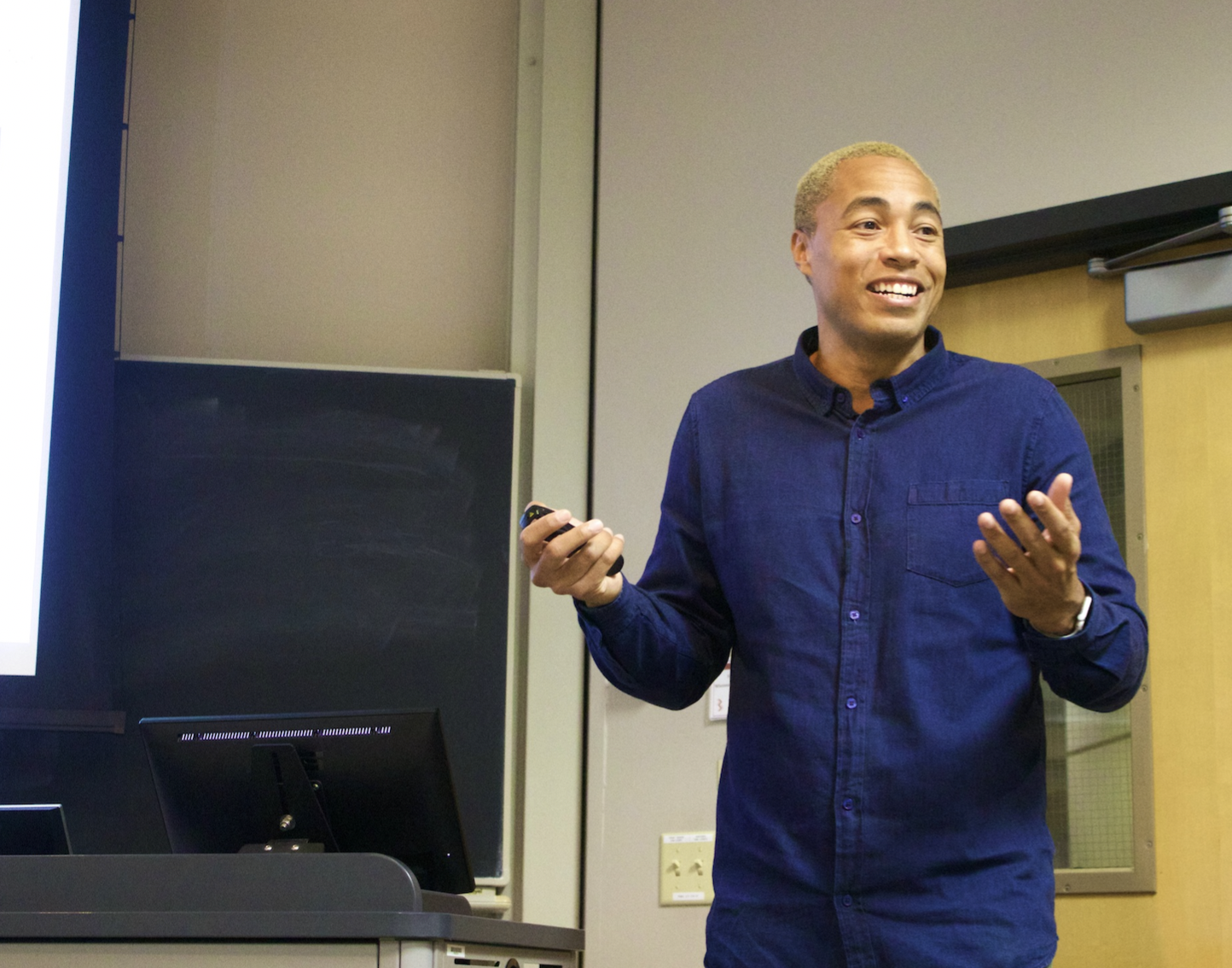 A man giving a presentation in a classroom, holding a remote control, smiling, wearing a navy blue shirt.