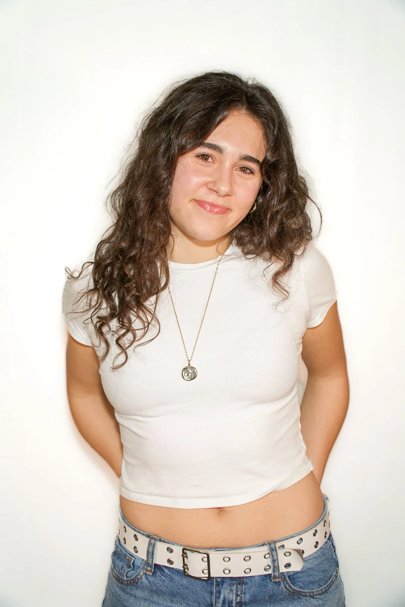 A young woman with long, curly dark hair wearing a white t-shirt, a necklace, and jeans with a white belt, standing against a plain white background.