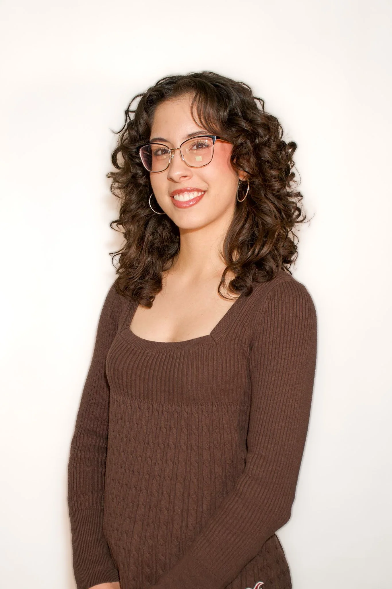 A young woman with curly brown hair, wearing glasses and hoop earrings, smiling while standing against a plain white background.