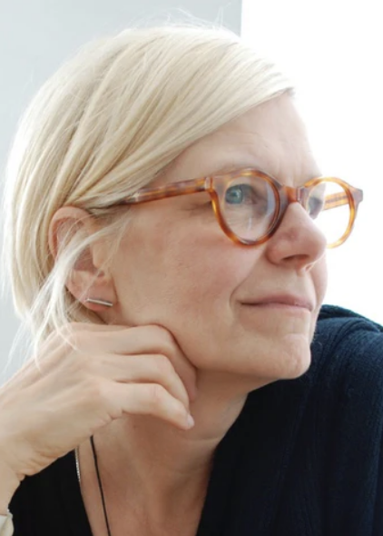 Close-up of a woman with blonde hair, wearing tortoiseshell glasses and a dark top, looking thoughtfully to the side with her hand near her face.