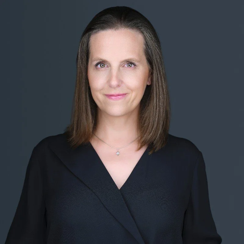 Young woman holding a black hat and bag, wearing a long, striped shirt and jeans, standing on a city sidewalk near a brick building.