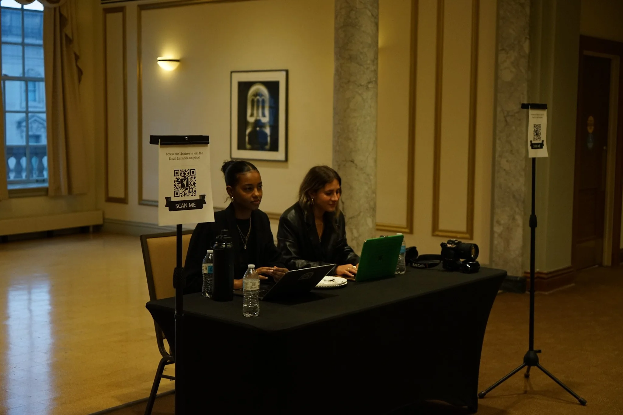 Two women sit at a black table with laptops and water bottles, inside a well-lit room with cream-colored walls and large windows. Sign with a QR code on a stand labeled 'Scan Me' is on the table.