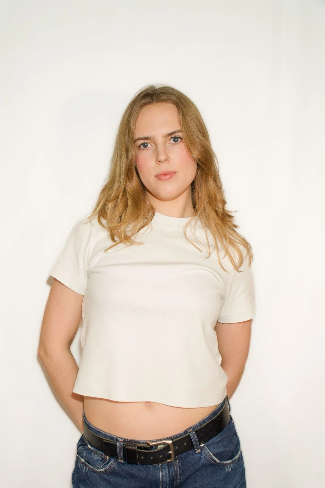 Young woman with blonde hair, wearing a white t-shirt and blue jeans, standing against a plain white background.