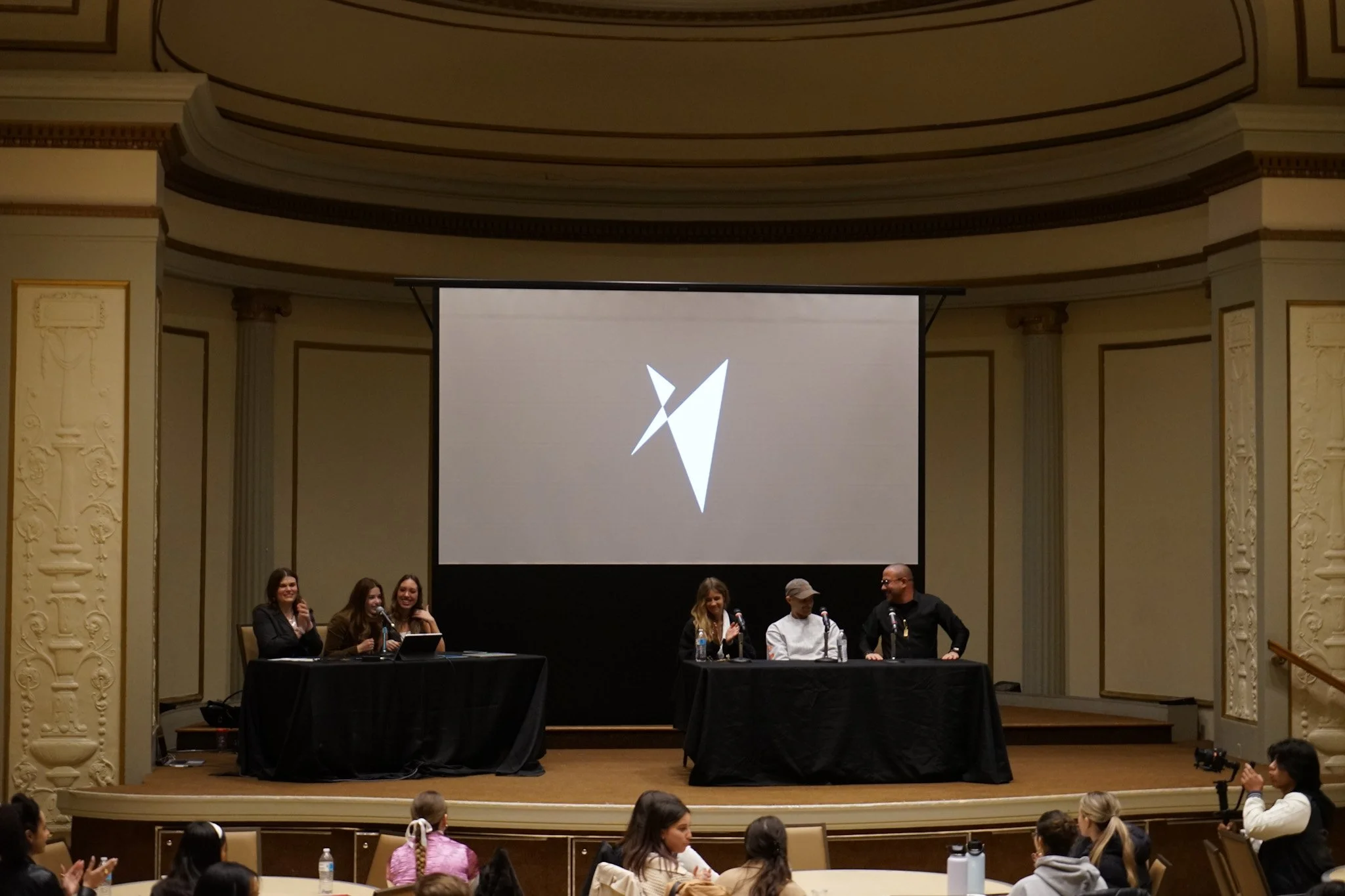 Panel discussion at a conference with five people on stage, three women and two men, seated at tables with microphones, in front of a large screen displaying a stylized logo. Audience members watch and applaud.