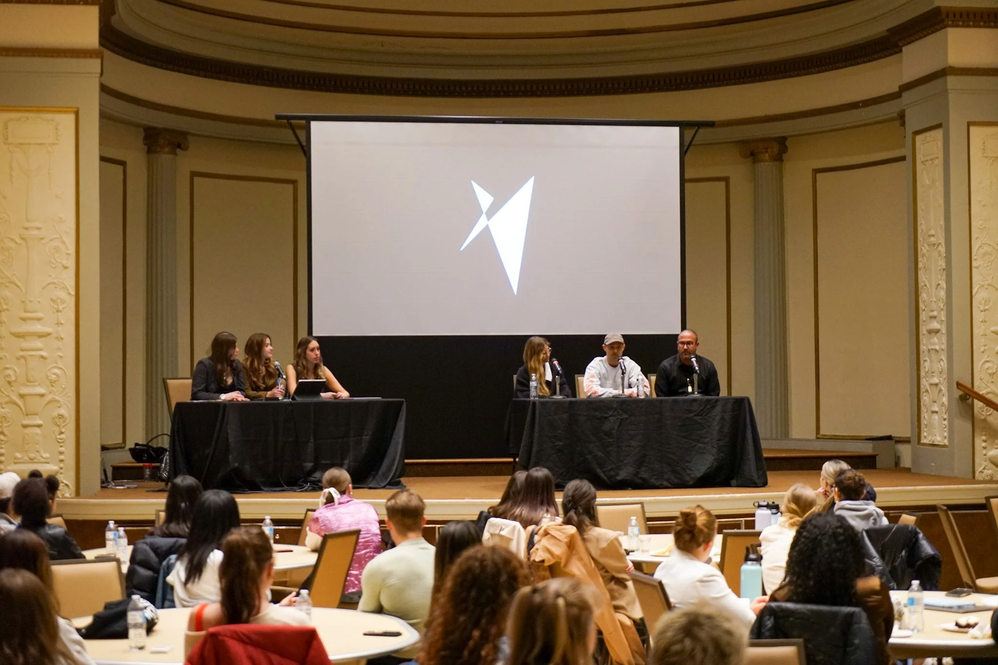 A panel of six people sitting at tables on stage in front of an audience, with a large blank screen behind them displaying a logo or graphic.