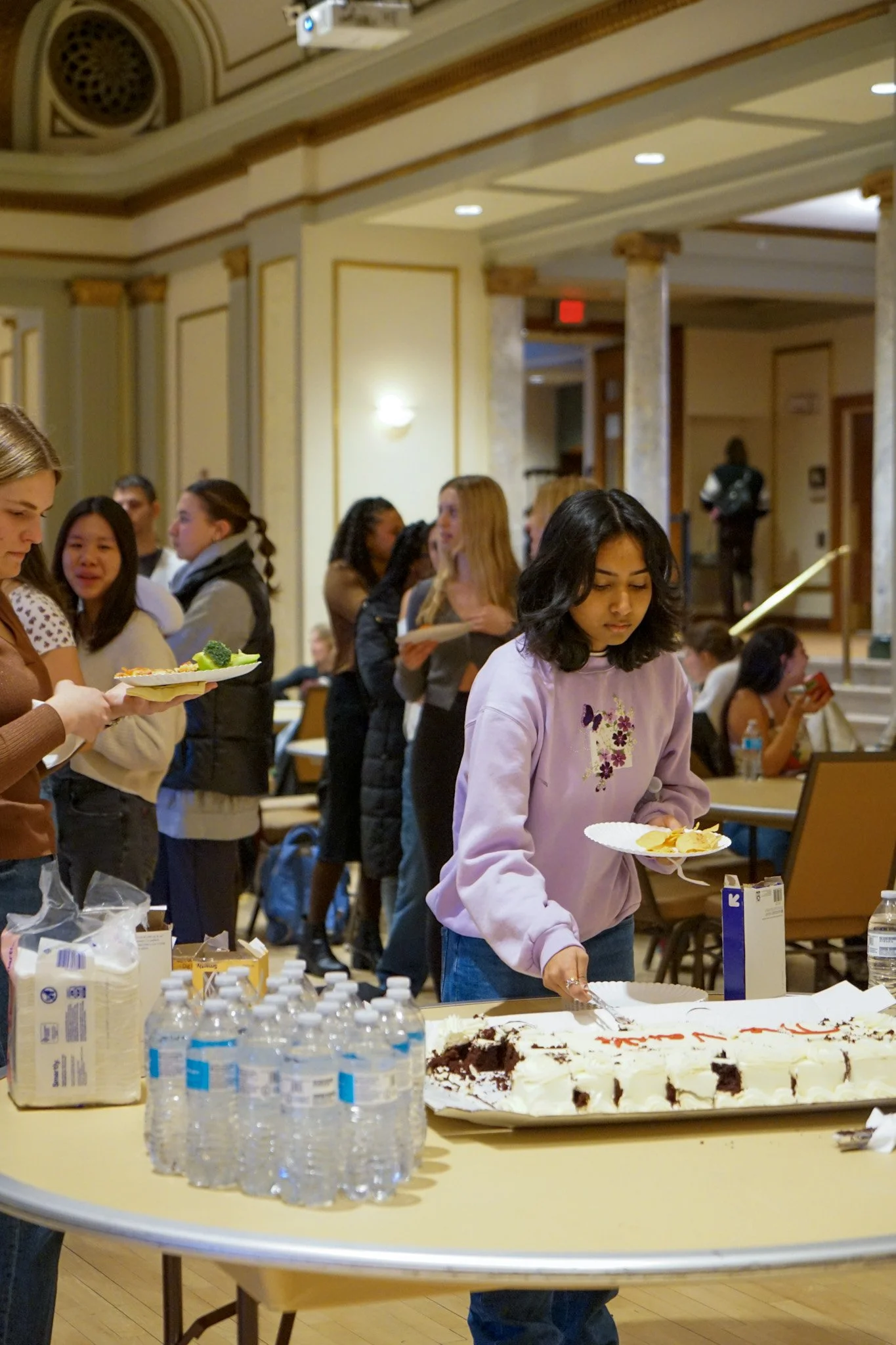 A group of young people at a social gathering, with some getting food and others waiting in line, in a room with ornate decor and a table with water bottles and a cake.