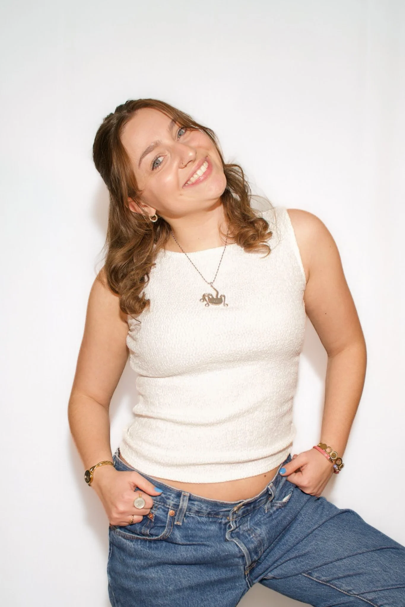 Young woman with brown wavy hair smiling, wearing a sleeveless white top, blue jeans, a necklace with a black panther pendant, and bracelets, standing against a plain white background.