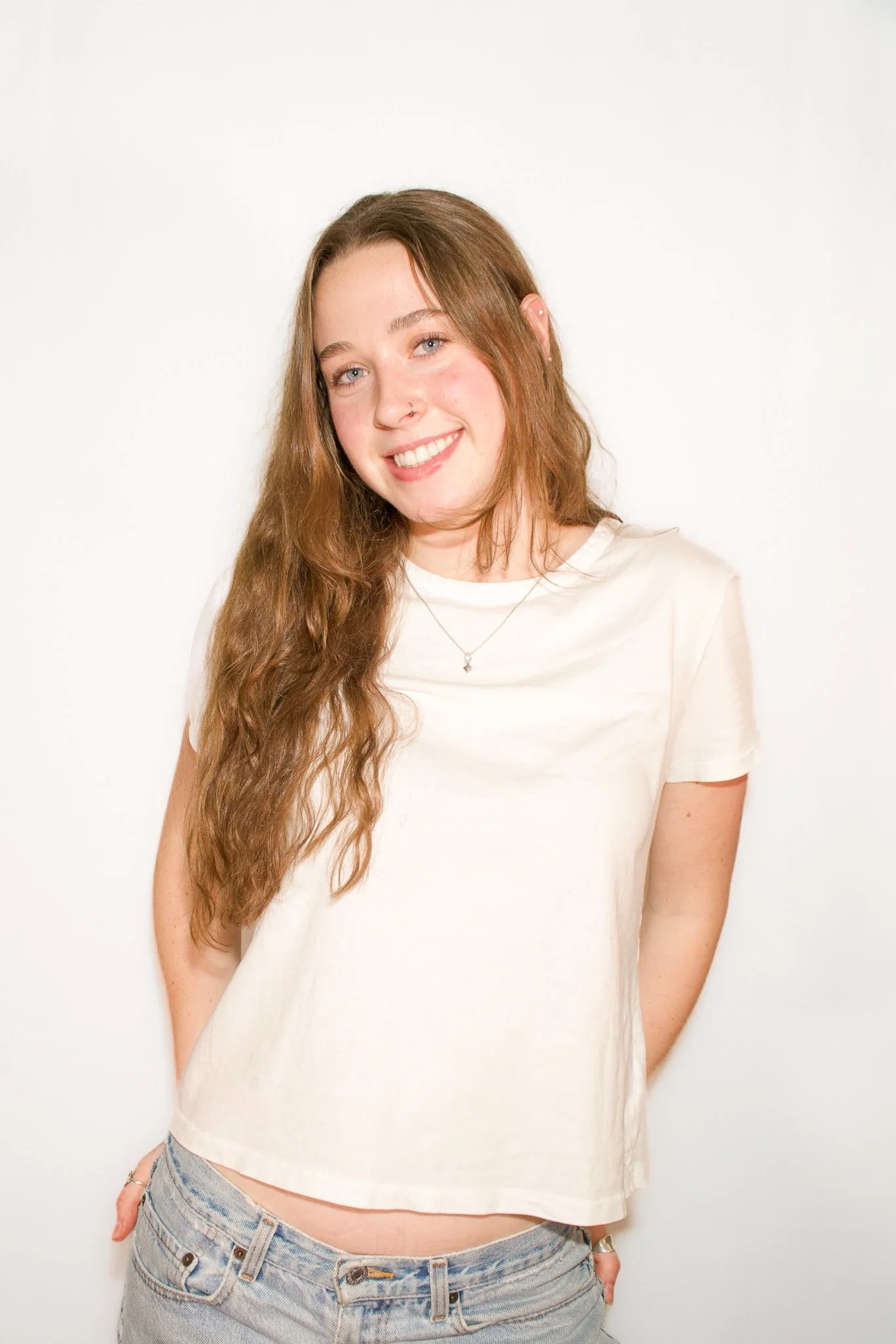 A young woman with long, wavy brown hair and light skin, smiling and looking at the camera, wearing a white t-shirt and light blue jeans, standing against a plain white background.