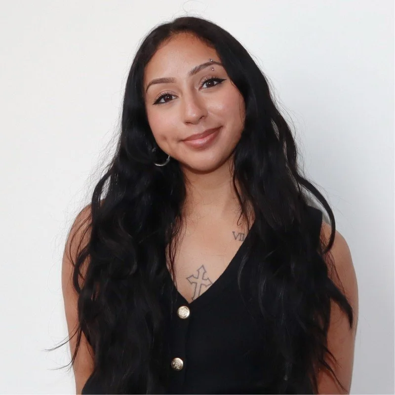 Young woman with dark hair in a bun, wearing a beige polka dot shirt, sitting with her chin resting on her hand, looking at the camera.
