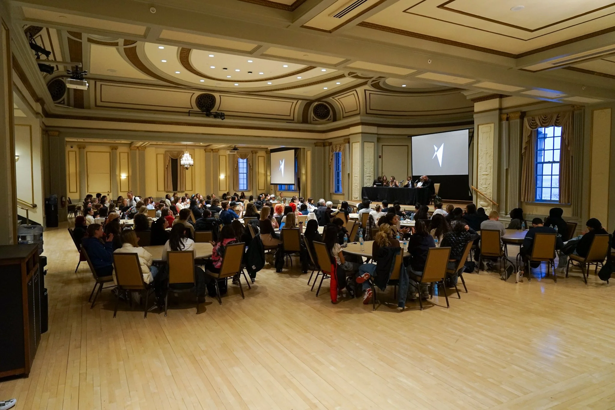 Large conference room filled with round tables and many people attending, with a stage at the front where a panel of speakers is seated, and two large projection screens displaying a logo.