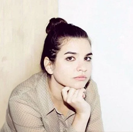 Young woman with dark hair in a bun, wearing a beige polka dot shirt, sitting with her chin resting on her hand, looking at the camera.