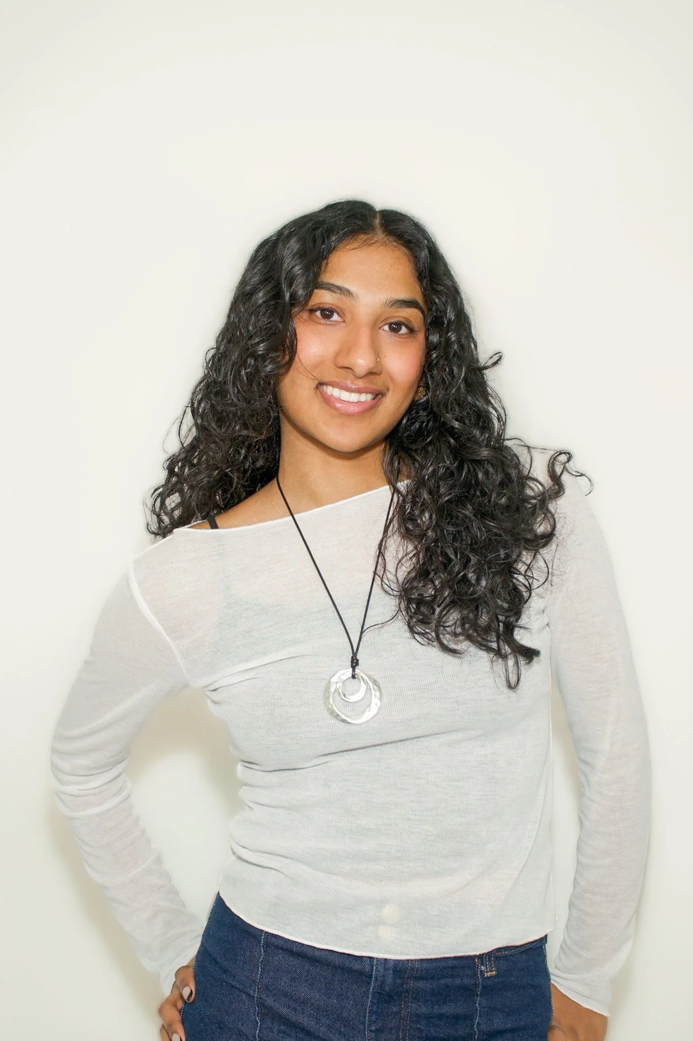 A young woman with curly black hair, wearing a white long-sleeve top and dark jeans, smiling at the camera against a plain white wall.
